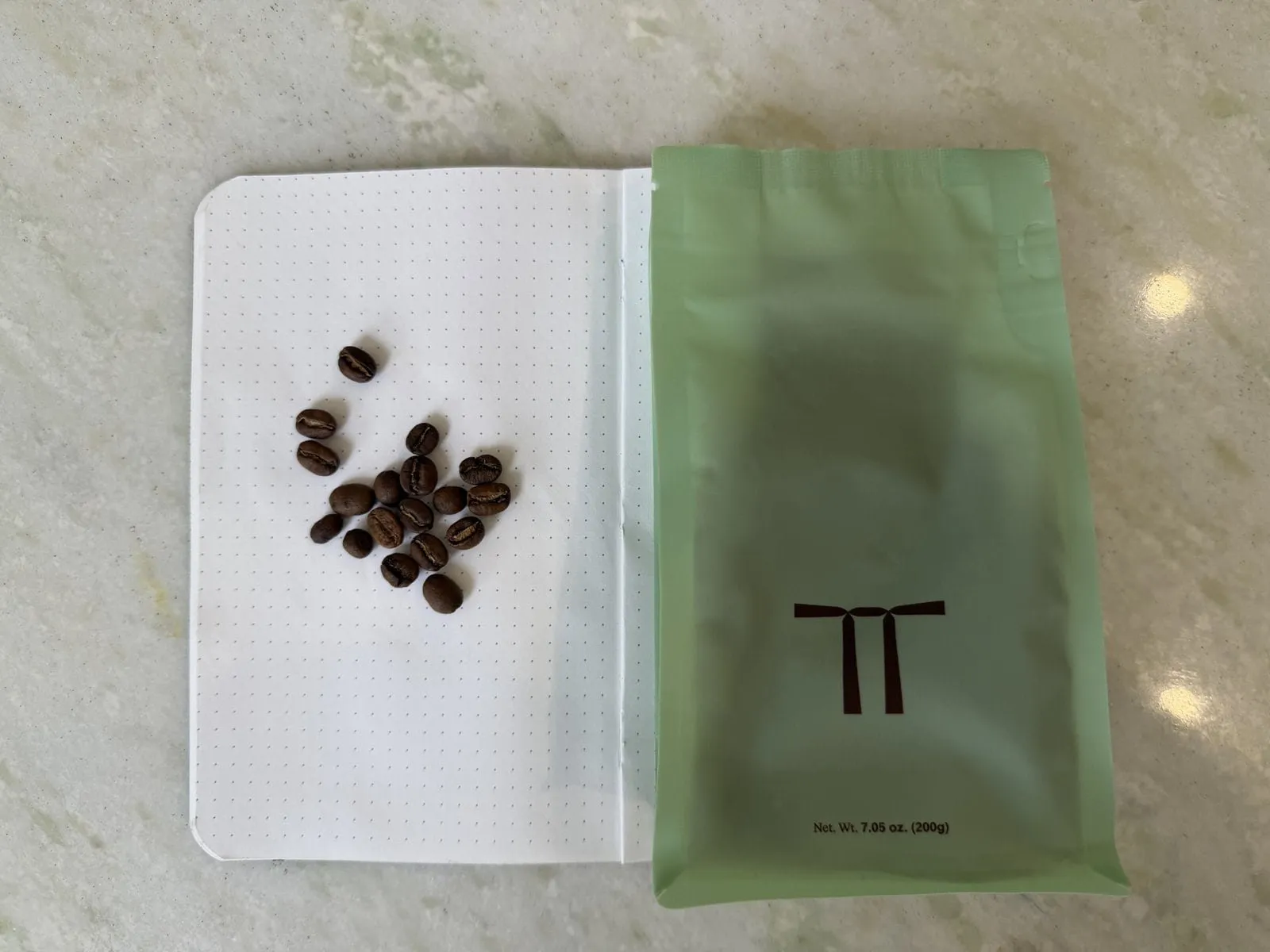 Coffee beans on a white dotted notebook page next to a green coffee bag with a minimalist logo.