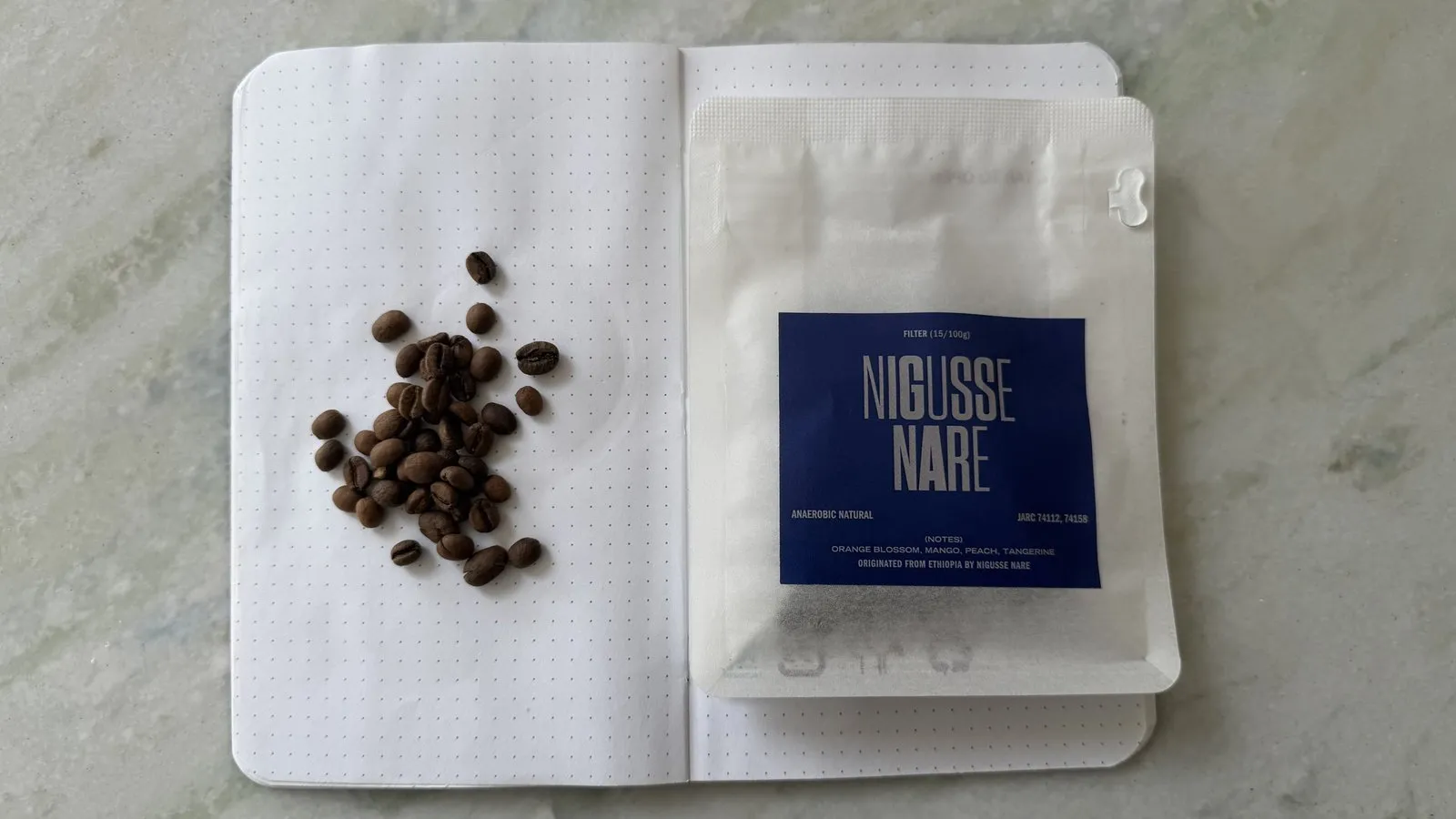 Close-up of NIGUSSE NARE coffee beans in a white notebook with a blue label package on a light grey surface.